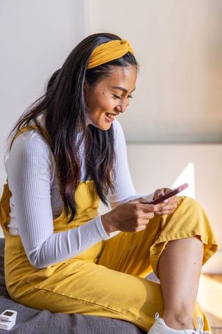 Smiling asian woman texting on smartphone while sitting on sofa in yellow overalls