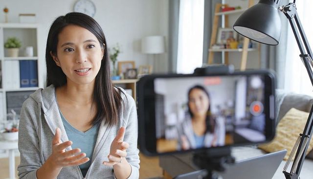 Asian woman recording casual vlog at home studio with smartphone on tripod, gesturing