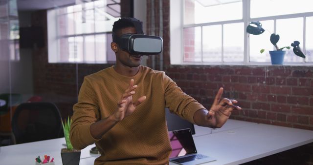 A young man wearing a VR headset is interacting with a virtual environment in a modern office with brick walls. The scene suggests innovative and futuristic activities, making it suitable for use in articles or advertisements related to technology, innovation, virtual reality advancements, and creative workspaces.