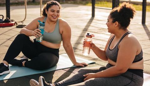 Smiling plus-size women resting and hydrating on outdoor exercise mats after workout
