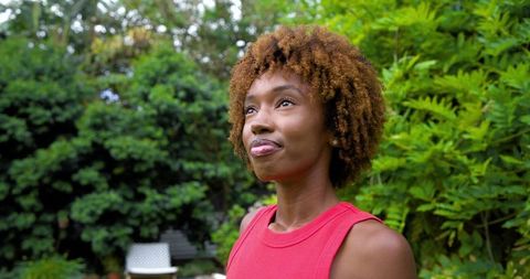 African american woman standing in garden looking reflective with curly hair red top