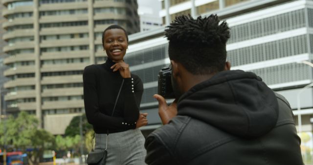A young African American woman smiles as she poses for a photo, taken by a photographer whose back is to the camera, with copy space. Capturing candid moments in an urban setting, the scene reflects a casual photoshoot or a moment of shared creativity between friends.