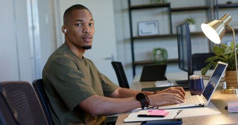 African american man typing on laptop while glancing at camera in modern shared workspace