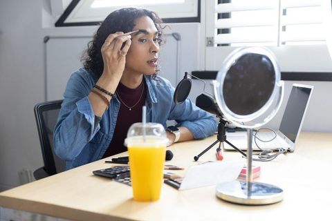 Non-binary creator grooming eyebrows at desk vanity with microphone, makeup, laptop