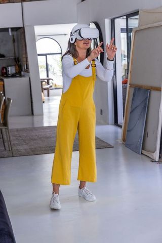 Middle-aged woman exploring virtual reality in bright home art studio in yellow overalls