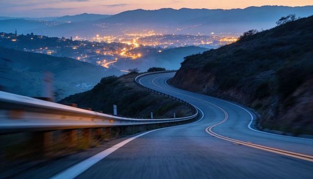 Twilight winding mountain road curving to lit valley with guardrail and city lights