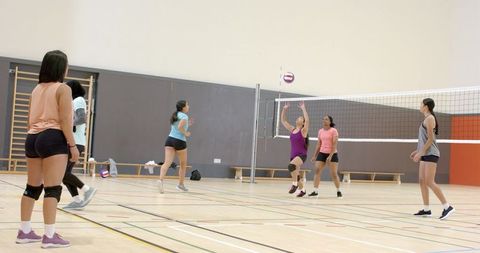 Diverse female volleyball team playing indoor gym jumping for ball during practice session