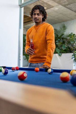 Indian man wearing orange sweatshirt playing pool holding red drink in modern lounge