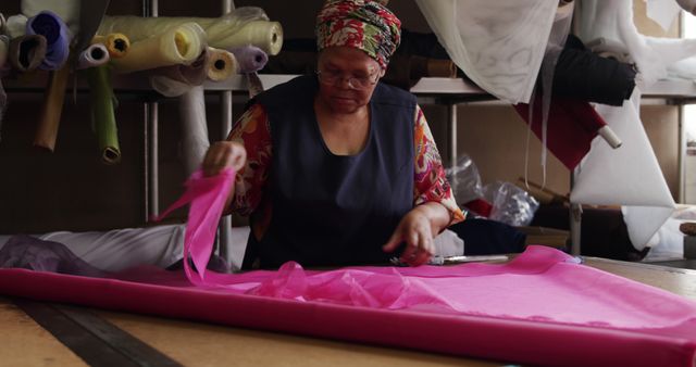 Senior seamstress working with pink fabric in a workshop, focusing on precision and detail as she prepares the material. Rolls of various fabrics and sewing equipment are visible in the background. Suitable for use in articles about craftsmanship, sewing professions, traditional skills, and handmade textile products. Ideal for advertisements or content highlighting textile industry or skilled artisans.