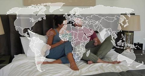 Couple relaxing on king bed with world map overlay, planning travel on phone and tablet