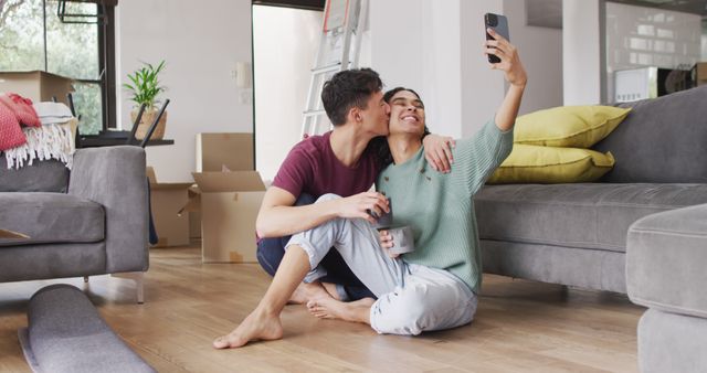 Couple sitting on the floor of their new home surrounded by moving boxes, taking a selfie together. It is a bright and clean modern living room with grey couches and colorful pillows. Both individuals look happy and excited, symbolizing a new beginning and the joys of moving into a new home. This image could be used for content related to real estate, moving services, relationship happiness, or home decor.