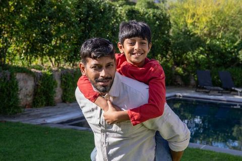 Joyful indian father and son playing piggyback by pool in sunny garden