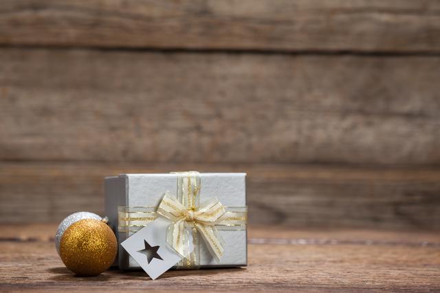 Wrapped gift box with ribbon and bow, accompanied by gold and silver baubles, on rustic wooden table. Ideal for holiday greeting cards, festive advertisements, Christmas promotions, and seasonal blog posts.