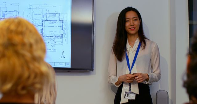 Asian businesswoman wearing a lanyard presenting to a diverse group of colleagues. A floor plan diagram is on the screen in the background. Perfect for topics related to leadership, diversity in the workplace, business meetings, professional growth, and teamwork.