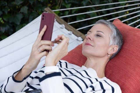 Mature woman reclining on hammock scrolling smartphone, enjoying relaxed garden serenity