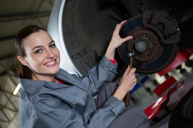 Female mechanic dressed in work clothes, smiling while working on car brake system in a well-lit auto garage. Useful for depicting gender diversity in automotive professions, car repair services, and promoting women in technical fields.