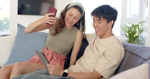 Young couple taking selfie while relaxing on couch and using tablet in bright living room