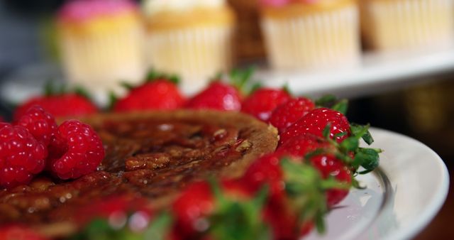 Pecan pie garnished with fresh raspberries and strawberries presents on a white plate. Blurred background features assorted cupcakes for added context. Ideal for use in recipes, dessert blogs, food magazines, bakery advertisements, and culinary websites highlighting homemade desserts.
