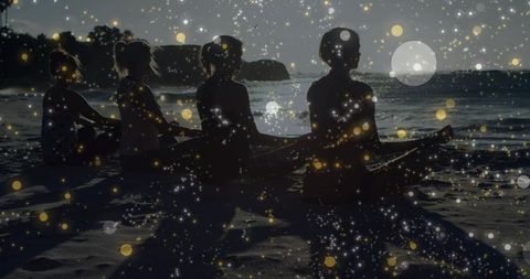 Silhouetted women sitting on beach at dusk watching sun over ocean with canoe and bokeh