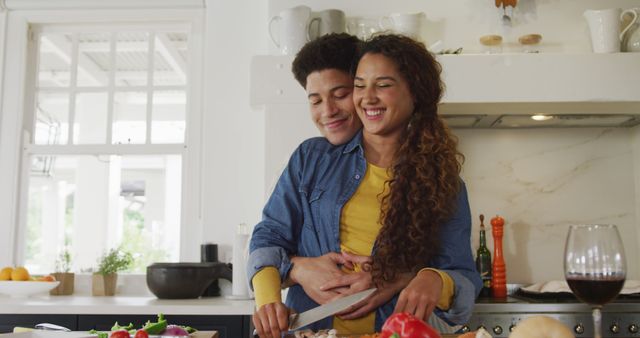 A young couple is cooking together in a bright kitchen, smiling and embracing each other. The scene exudes warmth, happiness, and affection, suggesting themes of love and domestic bliss. It can be used in articles about relationships, cooking tips, home life, or advertisements promoting culinary products, kitchen appliances, and healthy living.