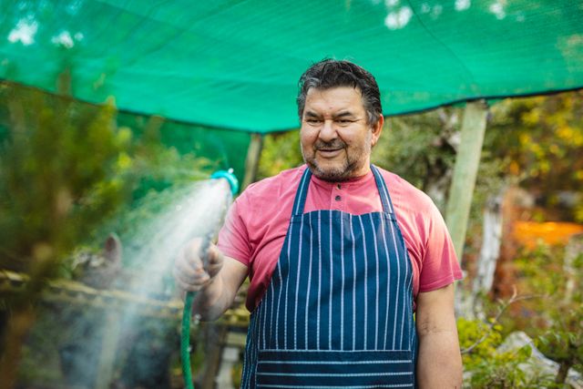 Male gardener wearing apron watering plants with hose in a garden centre. Ideal for use in articles about gardening, horticulture, small businesses, plant care, and outdoor activities. Can also be used in promotional materials for garden centres and nurseries.
