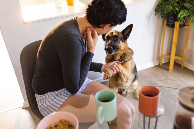 Lesbian woman shaking hands with her German Shepherd in a cozy home setting. Ideal for use in articles or advertisements about pet care, LGBTQ+ lifestyle, morning routines, and domestic life. Perfect for promoting products related to pets, coffee, or home living.