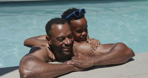 African american father and son enjoying sunny backyard pool, child hugging dad at edge