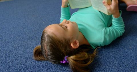 This scene illustrates a young girl lying comfortably on a blue carpet, focused on her book. She embodies the joy and tranquility of reading in a quiet school library. Her position suggests ease and adaptability to informal learning environments. This image is viable for educational materials, reading programs, library promotions, and articles about childhood literacy and personal development.