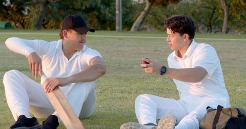 Two cricketers resting on grass exchanging bat and ball during late afternoon practice