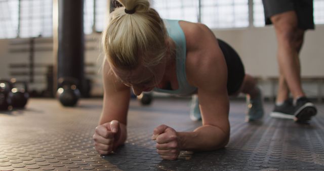 Blonde woman practicing plank exercise on gym floor, showing determination and focus. Useful for fitness blogs, workout programs, health and wellness websites, or gym advertisements related to core strength and personal training.