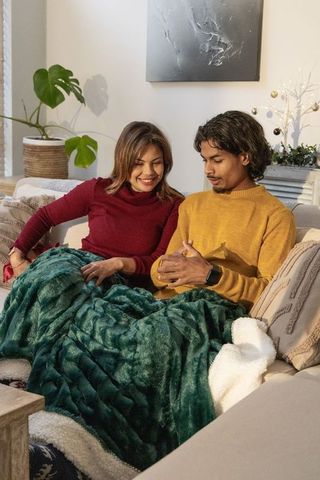 Hispanic woman and asian man relaxing on cozy sofa under green blanket using smartphone