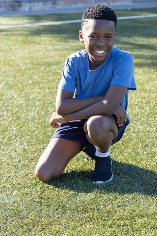 African american teen kneeling smiling on sunny sports field wearing blue shirt and shorts