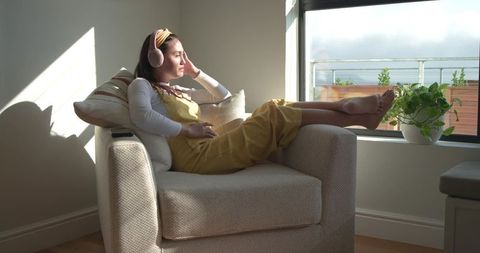 Mid-adult asian woman relaxing in armchair wearing yellow overalls and pink headphones