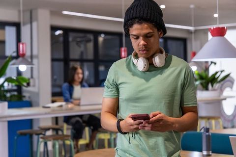 African american late-teen checking smartphone in modern coworking lounge with headphones