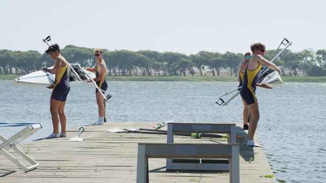 Capturing male rowers preparing their boats and oars by the lake on a sunny day. This scene is perfect for illustrating themes of teamwork, sport training, outdoor activities, and preparation. The peaceful lakeside setting combined with the active preparation of sportsmen adds a dynamic element to any visual project.