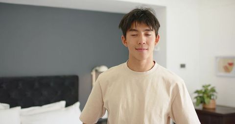 Young man standing relaxed in minimalist bedroom wearing beige t-shirt, calm lifestyle