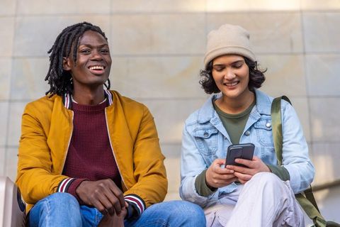 Friends laughing and checking smartphone together outdoors wearing casual denim and beanie