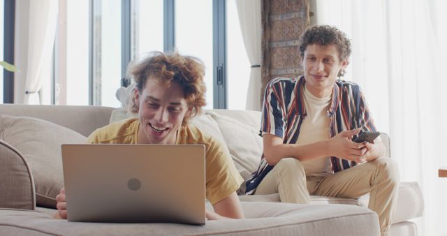 Happy diverse gay male couple sitting at sofa using tablet and smartphone at home, slow motion. Lifestyle, togetherness, communication and domestic life, unaltered.