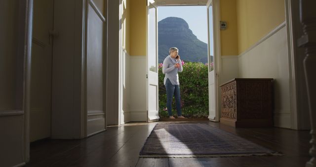 Hallway and senior caucasian woman by open doors in sunny garden talking on smartphone, copy space. Communication, mental health, domestic life and senior lifestyle, unaltered.