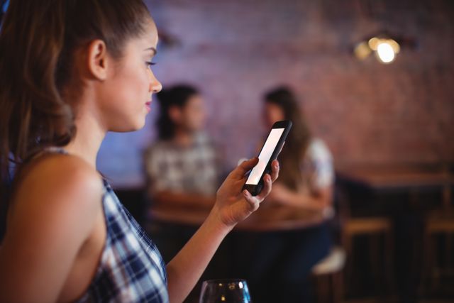 Young woman using mobile phone in pub, focusing on screen while others socialize in background. Ideal for themes related to technology, social media, nightlife, and modern lifestyle.