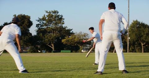 Cricket batsman playing red ball with teammates fielding on sunlit grassy ground