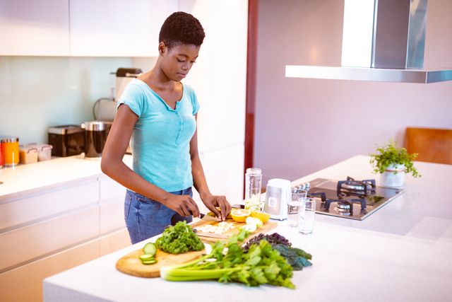 African American woman chopping fresh vegetables in a modern kitchen, preparing a healthy smoothie. Ideal for content related to healthy lifestyle, self care, wellness, cooking at home, and domestic life. Perfect for blogs, articles, and advertisements promoting healthy eating and home cooking.