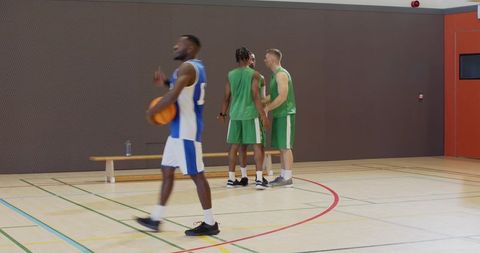 Basketball player walking with ball gesturing while teammates huddling on indoor court