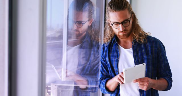 Modern professional with long hair and beard using digital tablet while leaning against reflective glass. Ideal for depicting modern work environments, tech industry roles, or casual business settings.