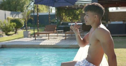 Young man lounging poolside on deck speaking into smartphone during sunny backyard leisure