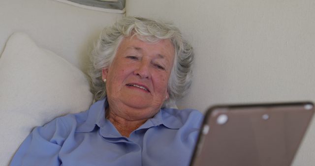 This image features an elderly woman comfortably reclining on a couch while using a digital tablet. Ideal for promoting technology usage among seniors, online education, digital communication, or products and services catering to the elderly.
