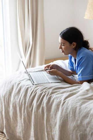 Female nurse lying on bed using laptop in bright bedroom working from home telehealth