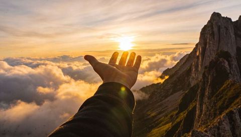Hand reaching toward golden sun above clouds on mountain ridge at sunset inspiration scene