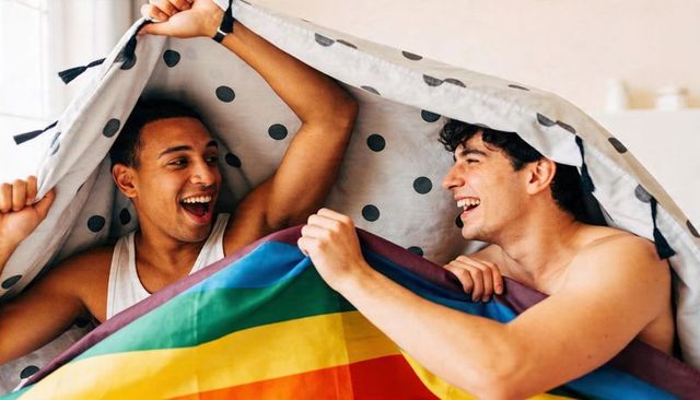 Joyful couple sharing rainbow flag under polka-dot duvet in cozy bedroom