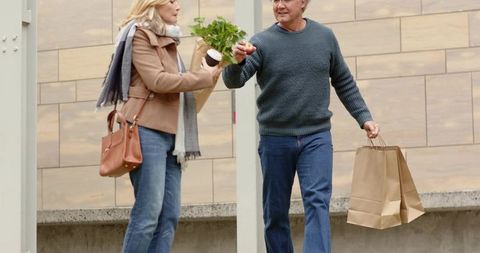 Mature couple walking through city plaza carrying groceries, coffee and paper bags in autumn
